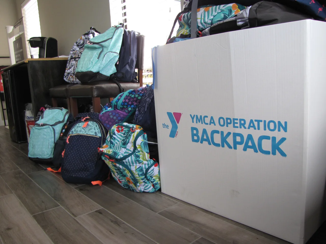 Rows of filled backpacks standing on a floor beside a large printed box reading YMCA Operation Backpack.