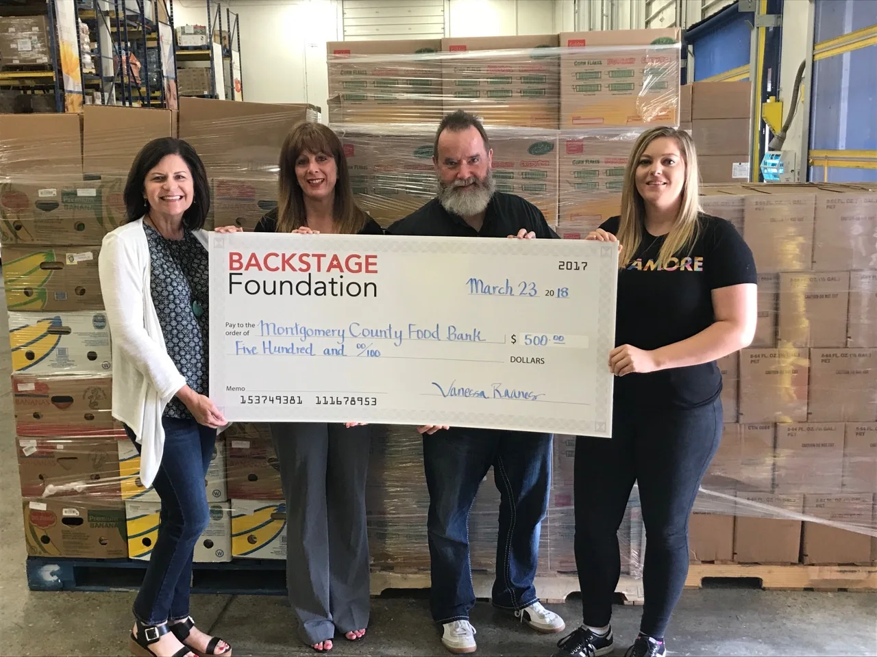 Four people standing in a food-bank warehouse holding a large presentation cheque from Backstage Foundation made out to Montgomery County Food Bank.